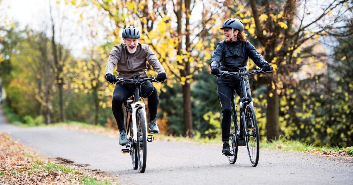 Smiling Senior Couple Riding E-Bikes on a Road in A Forested Area